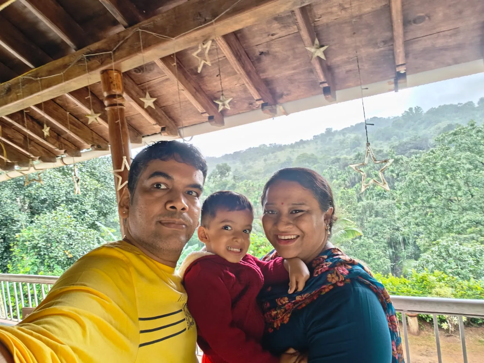 Phanindra with his wife and son on the balcony of the forest stay, with misty green hills behind.